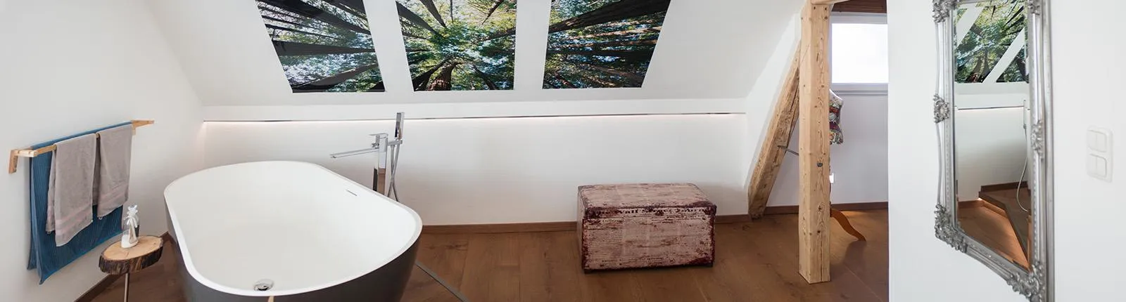Modernes Badezimmer im Dachgeschoss mit freistehender Badewanne, Holzfußboden, sichtbaren Holzbalken und großen Dachfenstern mit Naturmotiv; gemütliche Kombination aus Holz und Weißtönen.