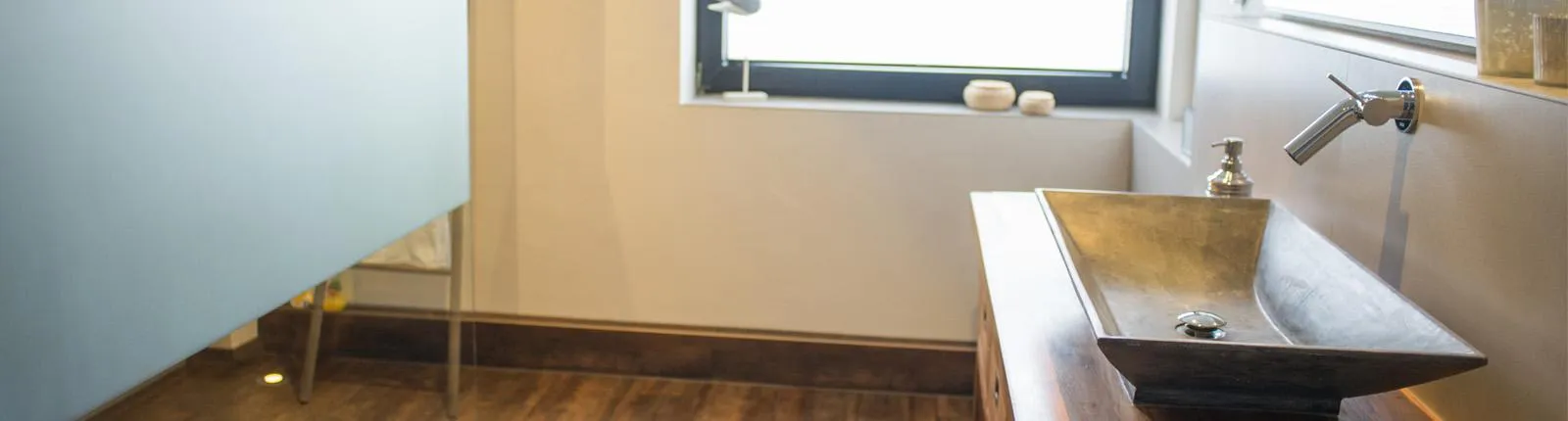 Moderner Waschtisch mit eckigem Steinwaschbecken auf Holzplatte, Wandarmatur aus Edelstahl, großem Fenster und Boden in Holzoptik.