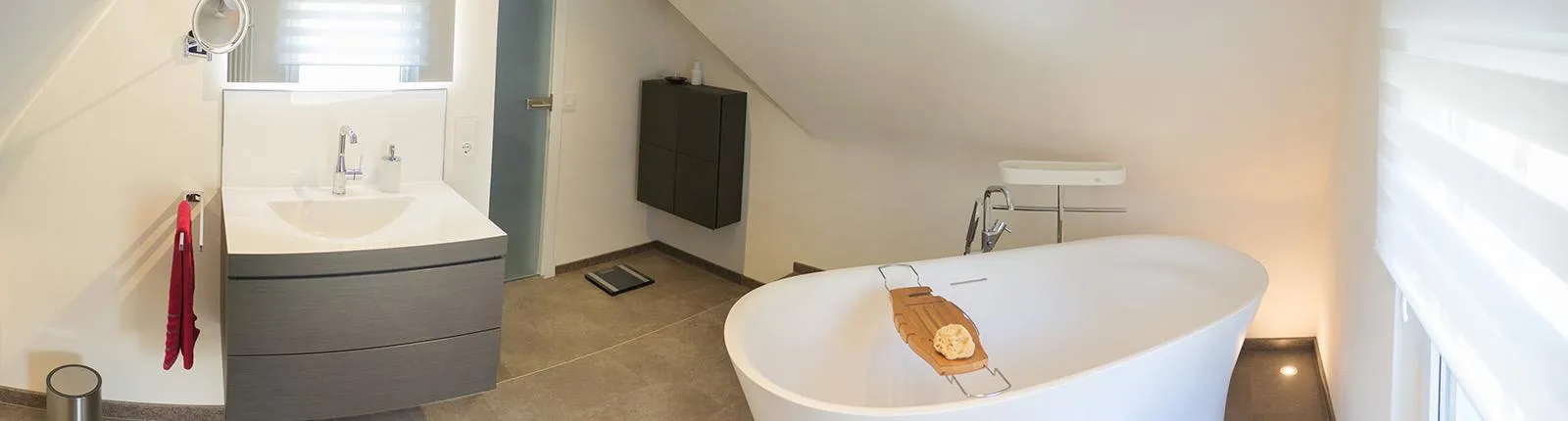 Modernes Badezimmer mit freistehender Badewanne, stilvollem Waschbecken, grauen Badmöbeln und schlichter Beleuchtung unter der Dachschräge.