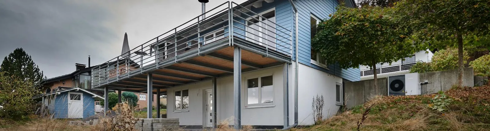 Modernes Wohnhaus mit blau-weißer Fassade, großer umlaufender Terrasse auf Stahlstützen, darunterliegendem Eingang, seitlich sichtbarer Wärmepumpen-Außeneinheit und umgebender begrünter Hanglage.