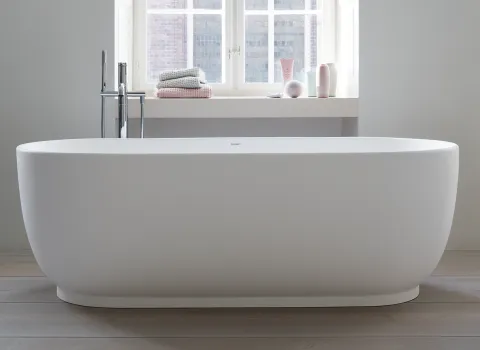 Eine freistehende, weiße Badewanne der Marke Duravit steht vor einem großen Fenster, durch das sanftes Tageslicht einfällt.