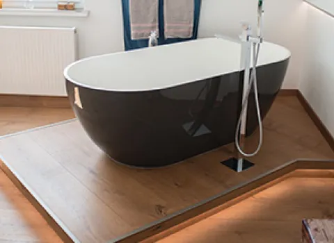 Freistehende schwarze Badewanne auf erhöhtem Holzpodest mit indirekter LED-Beleuchtung, moderner Armatur und Handtuchhalter im Hintergrund.