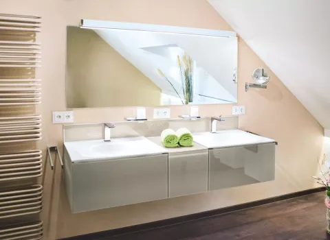 Modernes Badezimmer unter dem Dach mit Doppelwaschtisch, großem Spiegel, beigen Wänden, dunklem Boden und hellgrünen Handtüchern als Farbakzent.