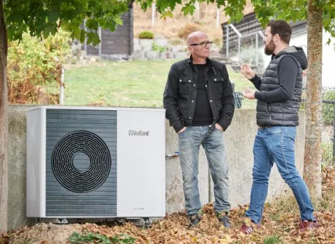 Zwei Personen stehen im Freien neben einer außen installierten Wärmepumpe von Vaillant und unterhalten sich, mit herbstlichen Blättern am Boden und Bäumen im Hintergrund.