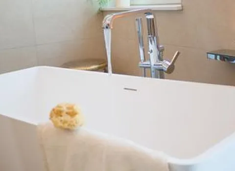 Weiße freistehende Badewanne mit moderner Standarmatur, aus deren Hahn Wasser fließt. Auf dem Wannenrand liegen ein Handtuch und ein Badeschwamm. Im Hintergrund sind beige Wandfliesen und ein kleines Fenster zu sehen, das Tageslicht hineinlässt.