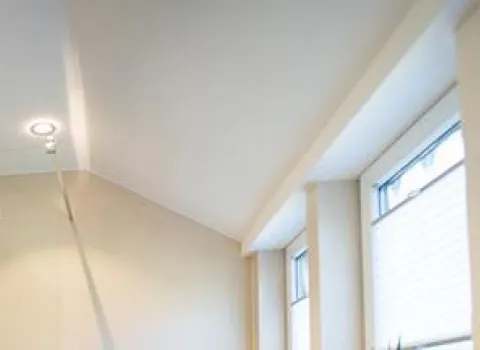 Ausschnitt eines modernen Badezimmers mit schräger weißer Decke, eingebautem Spotlicht und mehreren hohen Fenstern mit halb geschlossenen Jalousien, durch die Tageslicht einfällt.