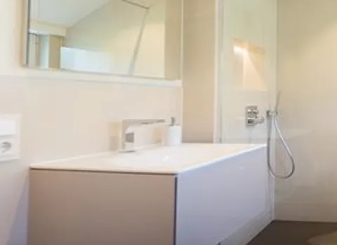 Ein modernes, helles Badezimmer mit einem weißen Waschtisch, großem Wandspiegel und einer offenen Dusche mit Handbrause.