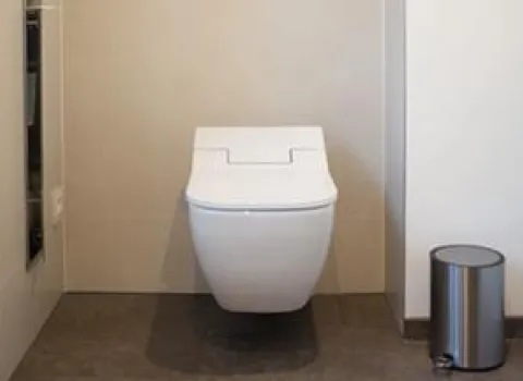 Eine moderne, wandhängende weiße Toilette in einem schlichten Badezimmer. Rechts daneben steht ein kleiner metallischer Mülleimer, der Boden ist dunkel und die Wände sind in neutralen Tönen gehalten.