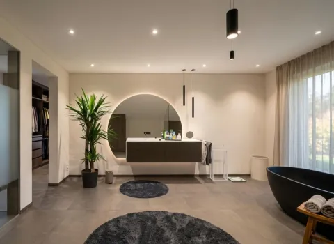 Modernes, luxuriöses Badezimmer mit freistehender schwarzer Badewanne, schwebendem Waschtisch, beleuchtetem Rundspiegel und Zimmerpflanze. Dunkle Bodenfliesen und der Zugang zu einem begehbaren Kleiderschrank ergänzen das minimalistische Design.