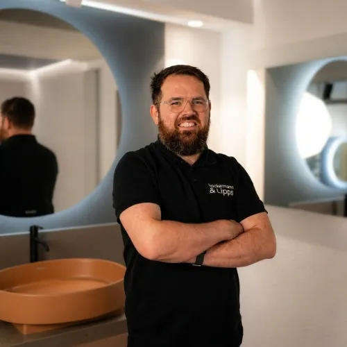 Ein lächelnder Mann mit Bart und Brille trägt ein schwarzes Poloshirt mit der Aufschrift „Ueckermann & Lipps“ und steht mit verschränkten Armen in einem modernen Badezimmer oder Showroom. Im Hintergrund sind ein graues ovales Waschbecken mit schwarzem Wasserhahn und mehrere runde sowie geschwungene Spiegel zu sehen.
