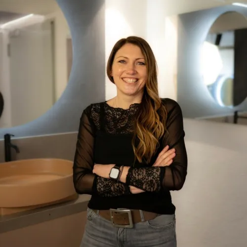 Eine lächelnde Frau mit langen, welligen braunen Haaren mit helleren Strähnen, trägt ein schwarzes Spitzenoberteil, graue Jeans, einen braunen Gürtel und eine Smartwatch. Sie hat die Arme verschränkt und steht in einem modernen Badezimmer mit einem ovalen, hellbraunen Waschbecken und kreisförmigen Spiegelelementen im Hintergrund.
