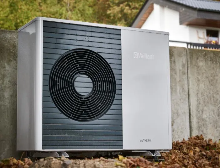 Außen aufgestellte Vaillant aroTHERM Wärmepumpe vor einer Betonmauer, umgeben von herbstlichem Laub, mit einem Wohnhaus im Hintergrund.