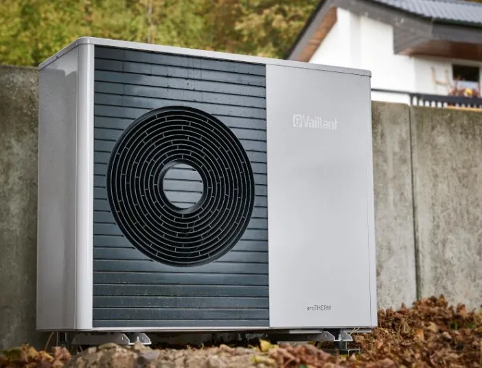 Außen aufgestellte Vaillant aroTHERM Wärmepumpe vor einer Betonmauer, umgeben von herbstlichem Laub, mit einem Wohnhaus im Hintergrund.