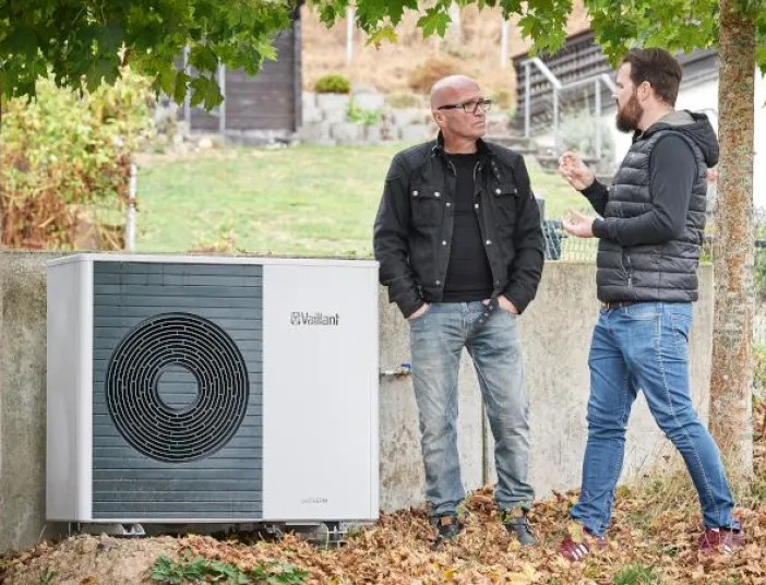 Zwei Personen stehen im Freien neben einer außen installierten Wärmepumpe von Vaillant und unterhalten sich, mit herbstlichen Blättern am Boden und Bäumen im Hintergrund.