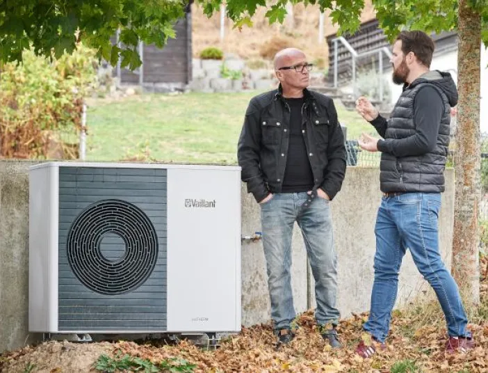 Zwei Personen stehen im Freien neben einer außen installierten Wärmepumpe von Vaillant und unterhalten sich, mit herbstlichen Blättern am Boden und Bäumen im Hintergrund.
