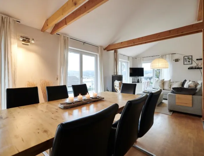 Helles, offen gestaltetes Wohn- und Esszimmer mit sichtbaren Holzbalken, großem Holzesstisch mit schwarzen Stühlen, Kerzenarrangement auf dem Tisch, bodentiefen Fenstern mit hellen Vorhängen, gemütlicher Sitzecke mit Sofa und Stehlampe sowie modernem Fernseher vor weiter Aussicht.