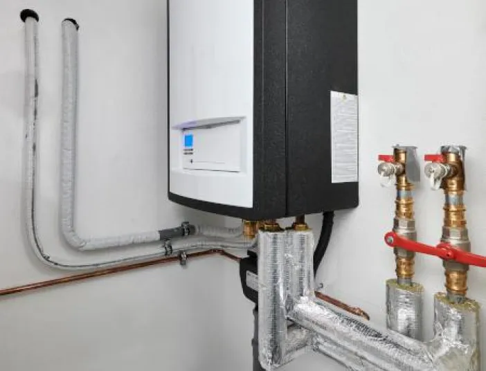 Wandmontierte Heizungsanlage mit weißem Frontpanel und schwarzem Gehäuse in einem Technikraum, angeschlossen an mehrere isolierte Rohrleitungen und Ventile, die entlang der Wand geführt sind.
