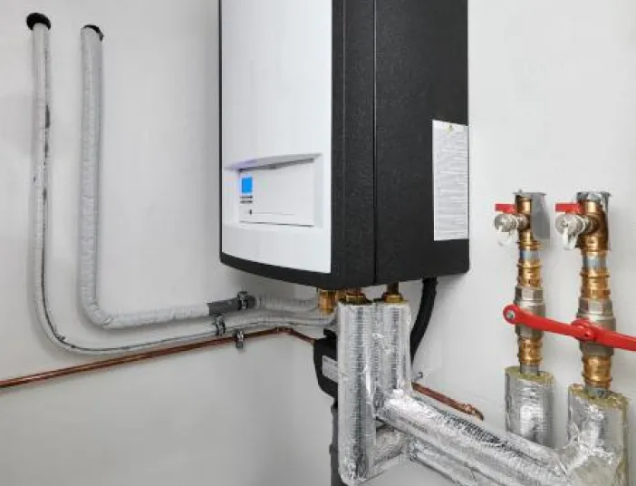Wandmontierte Heizungsanlage mit weißem Frontpanel und schwarzem Gehäuse in einem Technikraum, angeschlossen an mehrere isolierte Rohrleitungen und Ventile, die entlang der Wand geführt sind.