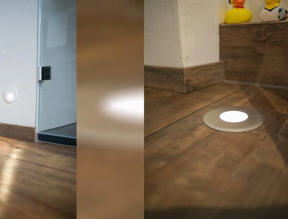 Detailaufnahme eines Badezimmers mit Holzoptikboden, integrierter LED-Bodenbeleuchtung und Wandlicht neben Dusche und WC.