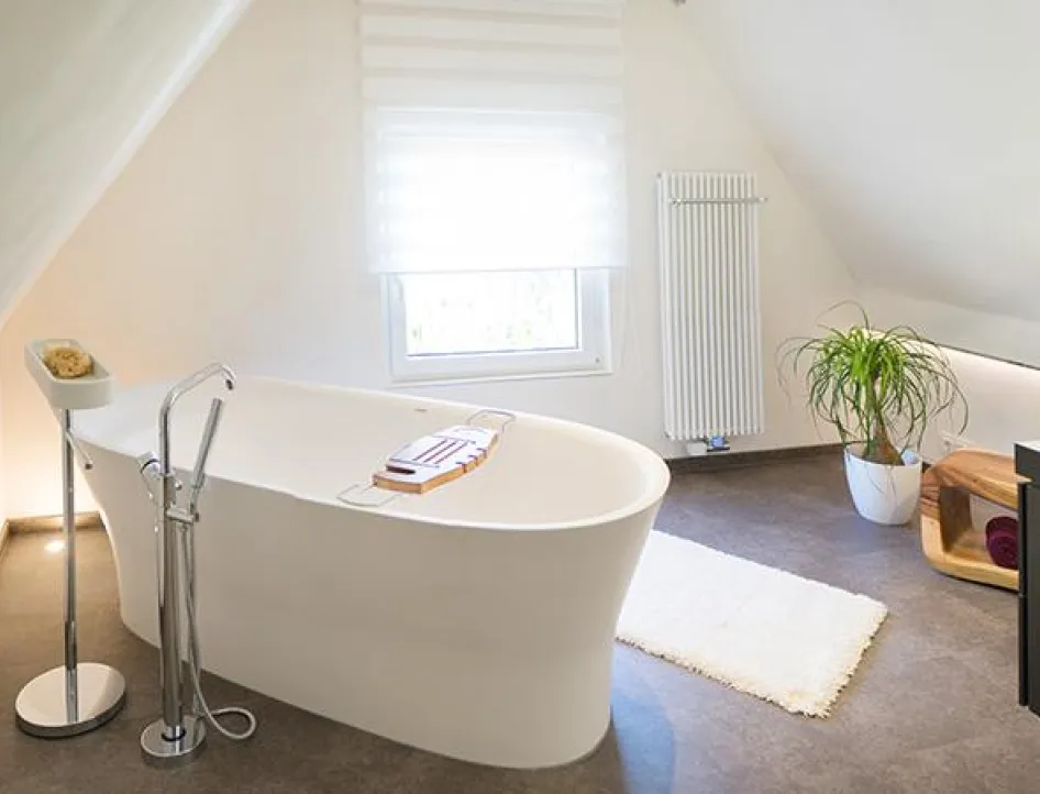 Helles, modernes Badezimmer mit freistehender Badewanne unter der Dachschräge, weißem Teppich, Zimmerpflanze und schlichten Holzelementen für eine ruhige Atmosphäre.